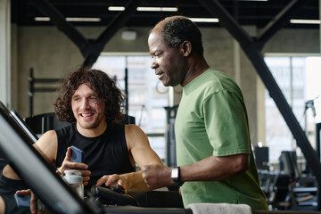 Fototapeta na wymiar Elderly african american man and caucasian athlete checking information on treadmill screen at gym