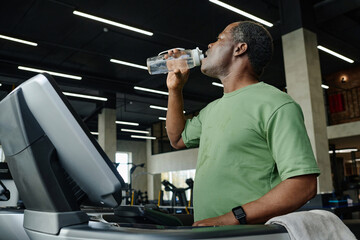 Medium shot of elderly black sportsman drinking water from bottle standing on treadmill at gym