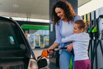 A mother pours fuel into the car while her daughter hugs her, both are happy