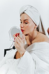Beautiful young applying red lipstick pencil, holding makeup mirror, towel wrapped on head