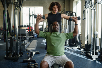 Senior african american man lifting dumbbells with young caucasian trainer helping him