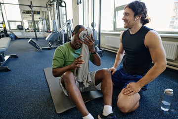 Senior black man sitting on floor at gym wiping forehead, caucasian instructor sitting next to him