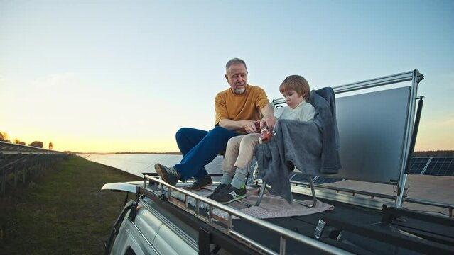 Grandfather and grandson resting atop camper roof, enjoying conversation within a vast solar power plant