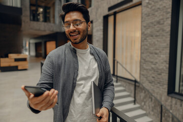 Smiling male manager in eyeglasses looking on mobile phone standing in modern coworking space 