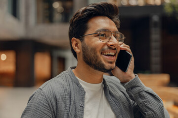 Smiling male manager in eyeglasses is talking phone standing in modern coworking space