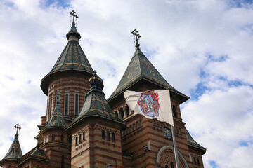 Fototapeta na wymiar The flag of Timisoara city waving against cloudy sky and Orthodox Cathedral Three Holy Hierarchs. Travel to Romania.