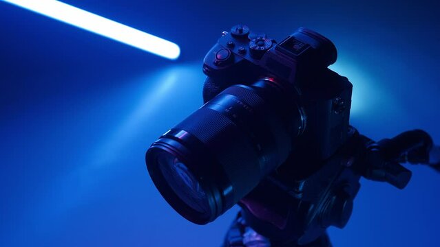 Professional photo camera stands on a tripod in a studio with color neon lighting.