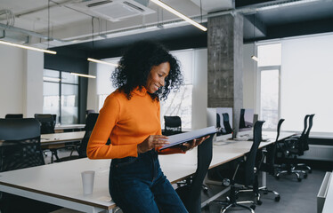 Smiling Black woman in an orange sweater reviews documents in a well-lit, modern office space
