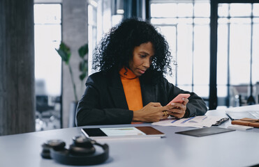 Diligent Black businesswoman in 30s, wearing gray blazer and orange turtleneck, engaged with smartphone