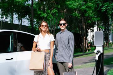 Young couple travel with EV electric car charging in green sustainable city outdoor garden in...
