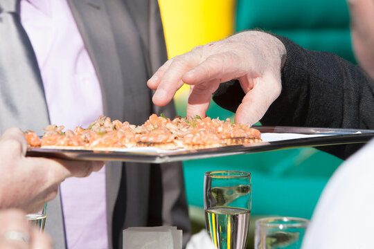 The image captures a moment at a catered event, with a server presenting a tray of smoked salmon canapes. An attendee is in the process of picking one of the delicately prepared appetizers
