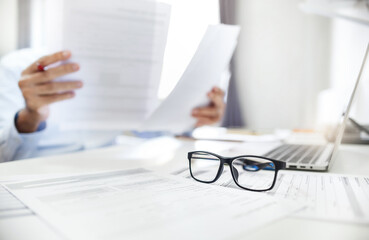 Close-up shot at eyeglasses, blurred businessman review many documents placed on the desk and using...