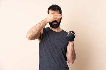 Caucasian sport man with beard making weightlifting over isolated background covering eyes by...