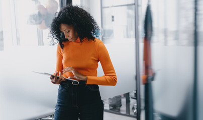 Intently focused Black businesswoman in an orange sweater navigating a tablet within a modern