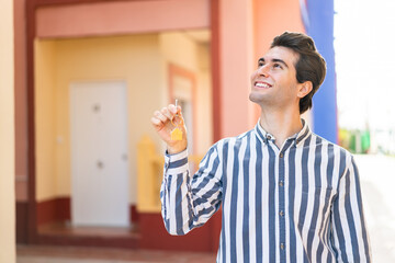 Young handsome man holding home keys at outdoors looking up while smiling