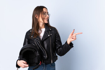 Young woman holding a motorcycle helmet over isolated blue background pointing finger to the side