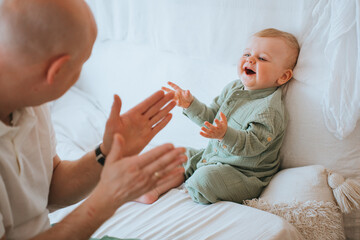 young dad claps his hands, plays with a baby boy on the bed, amuses him and makes him laugh....