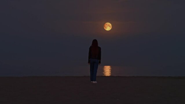 Moving shot of woman silhouette looking at the full moon light reflection on the surface of the water on the beach 