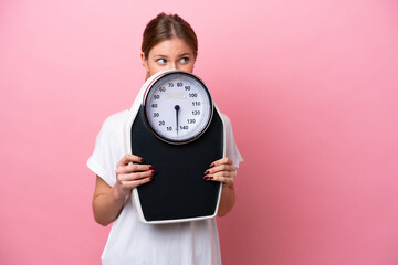 Young caucasian woman isolated on pink background with weighing machine and hiding behind it - obrazy, fototapety, plakaty
