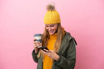 Young blonde woman wearing winter jacket isolated on pink background holding coffee to take away and a mobile - obrazy, fototapety, plakaty