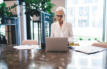 Fototapeta na wymiar Focused elderly woman working on laptop in modern meeting room
