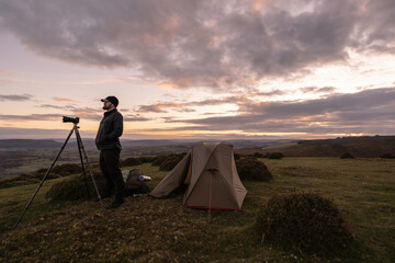 A wild camping explorer on the Shropshire Hills in England, stood beside his tent and camera tripod