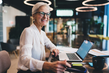 Happy aged woman in formal wear using laptop at workplace
