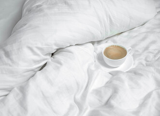 A white cup of coffee stands on a bed in white linen