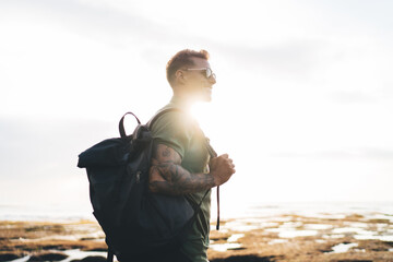 Happy man carrying backpack standing against ocean and sunlight - obrazy, fototapety, plakaty