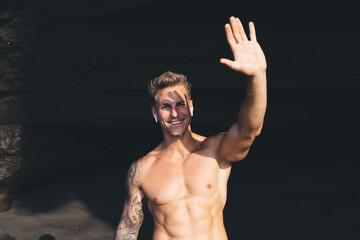 Fototapeta na wymiar Cheerful young man with naked torso raising hand while blocking sunlight