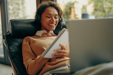 Young smiling female student studying on laptop and making notes while lying on chair at home
