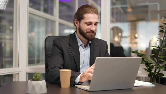 Smiling entrepreneur with trendy haircut typing email on his portable laptop during working day. Caucasian formally dressed male expressing positive emotion during inspiration with new idea.