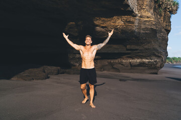 Obraz premium Shirtless man with raised arms on sandy beach by rocks