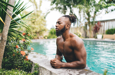 Confident shirtless black man standing in pool while resting hands on poolside