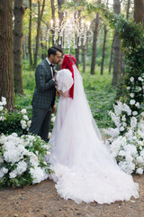 Beautiful couple, the groom in a green suit and the bride in an unusual pink wedding dress, posing at a luxurious ceremony with beautiful decor and an arch with a chandelier and candles in the forest.