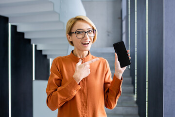 Portrait of a young woman standing in the office and enthusiastically pointing at the mobile phone...