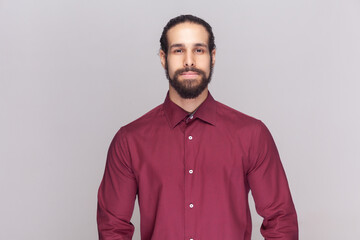 Portrait of serious strict bossy man with dark hair and beard in red shirt standing looking at camera, having confident facial expression. Indoor studio shot isolated on gray background. - obrazy, fototapety, plakaty