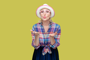 Portrait of senior woman wearing checkered shirt, hat and eyeglasses stretches hands near face, blows air kiss, expresses love and affection. Indoor studio shot isolated on yellow background.
