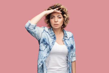 Portrait of serious attentive woman with curly hairstyle wearing blue shirt standing with palm near forehead, looking, trying to see something. Indoor studio shot isolated on pink background.