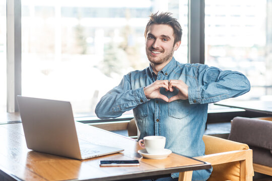 Portrait of smiling romantic bearded young man freelancer in blue jeans shirt working on laptop, showing heart shape, love gesture to camera. Indoor shot near big window, cafe background.