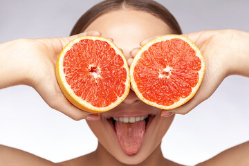 Closeup portrait of funny cheerful childish beautiful woman covering eyes with grapefruits showing tongue out. Indoor studio shot isolated over gray background.