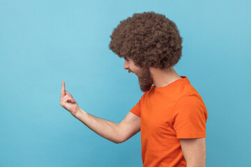 Side view of angry man with Afro hairstyle in orange T-shirt showing middle finger and asking to get off expressing negativity, disrespectful behaviour. Indoor studio shot isolated on blue background.