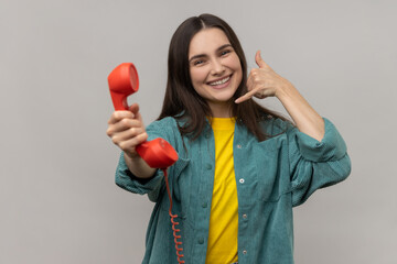 Beautiful smiling woman holding red headset of retro landline telephone and showing call me gesture, call center, wearing casual style jacket. Indoor studio shot isolated on gray background.