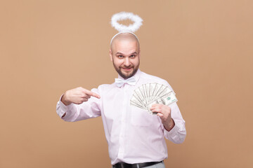Portrait of rich bearded man with nimb over head, holding and pointing at dollar banknotes, looking at camera, wearing light pink shirt and bow tie. Indoor studio shot isolated on brown background.