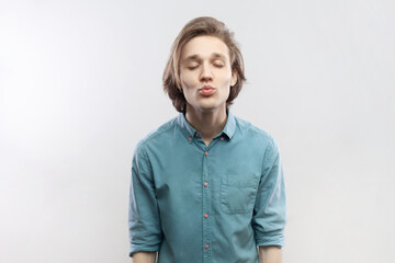 Obraz na płótnie Canvas Portrait of cute charming romantic young man standing with closed eyes and pout lips, sending air kisses, flirting with girlfriend, wearing blue shirt. Indoor studio shot isolated on gray background.
