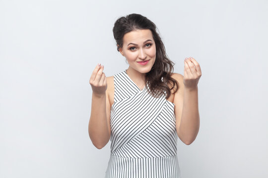 Portrait of woman outmakes money gesture, thinks about profitable business, demonstrates symbol of currency and richness, wearing striped dress. Indoor studio shot isolated on gray background.