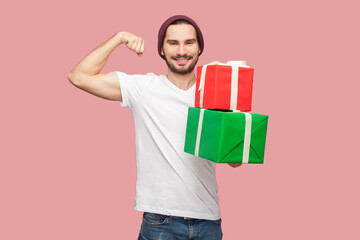 Portrait of smiling optimistic bearded man in white T-shirt and beany hat standing looking at camera, holding present boxes, showing biceps, raised arms. Indoor studio shot isolated on pink background