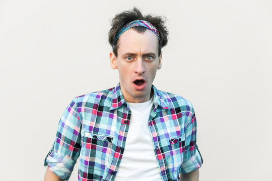 Stupefied man looks with astonishment at camera, being amazed with negative news, doesn`t believe his eyes, wearing checkered shirt and headband. Indoor studio shot isolated on gray background.