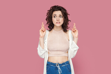 Portrait of hopeful attractive woman with curly hair wearing casual style outfit crossing her finger for good luck, waiting for miracle. Indoor studio shot isolated on pink background. - obrazy, fototapety, plakaty