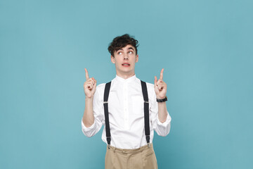 Portrait of confused attractive young man wearing white shirt and suspender, pointing finger up, showing advertisement area, copy space. Indoor studio shot isolated on blue background.